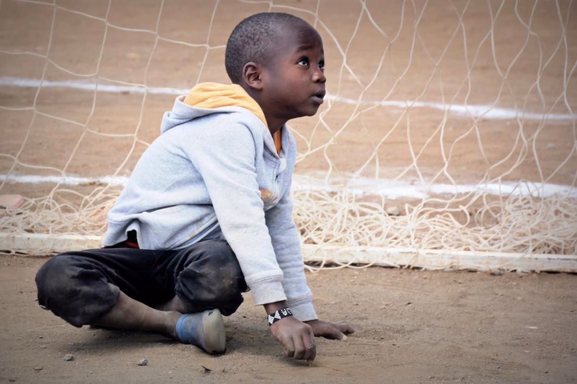 Ein Kind aus dem Slum Korogocho in Nairobi, Kenia, sitzt hinter einem Fußballtor auf dem Boden und schaut nach oben. | Foto © Tom Rübenach